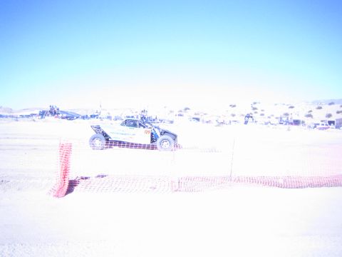 1990 Ford Bronco - dark off-road race buggy runs a sandy desert course behind orange safety fencing with distant pit vehicles.