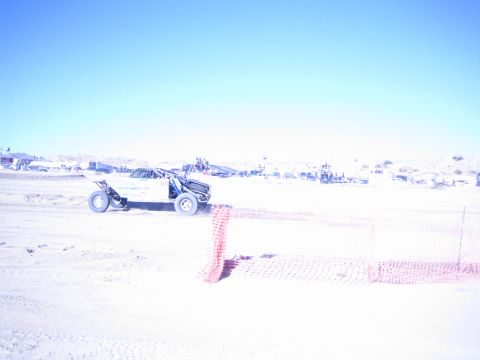 1990 Ford Bronco - dark off-road race buggy with a roll cage runs a sandy desert course behind orange fencing with distant pits.