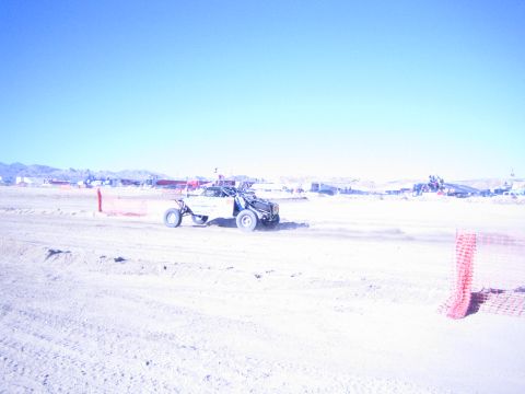 1990 Ford Bronco - off-road race buggy crosses a wide sandy desert course between orange fencing with distant pits and mountains.