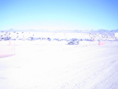 1990 Ford Bronco - lone off-road race buggy crosses a sun-bleached desert course with orange fencing and pit area in the distance.