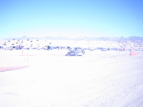1990 Ford Bronco - light-bodied off-road race car runs a desert course past orange fencing and a line of pit structures.