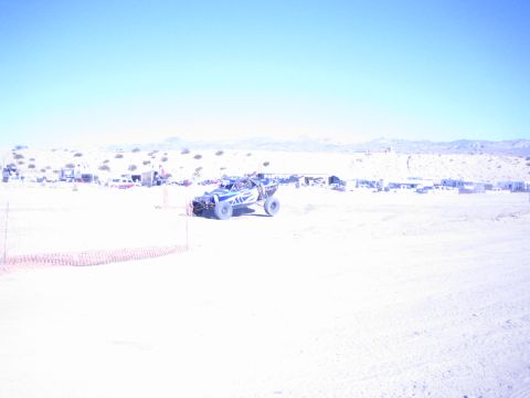 1990 Ford Bronco - off-road race truck pitches up over a rise on a desert course with orange safety fencing alongside.