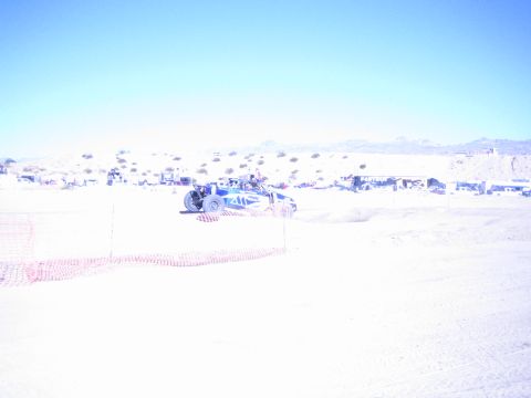 1990 Ford Bronco - blue off-road buggy runs a desert race course behind orange snow-fencing with pits and mountains beyond.