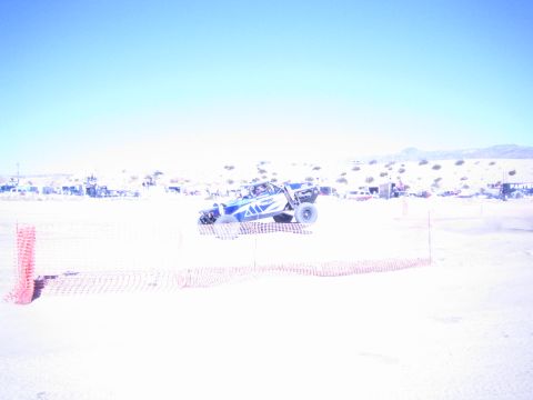 1990 Ford Bronco - blue-and-white off-road buggy catches air over a desert race course lined with orange fencing.