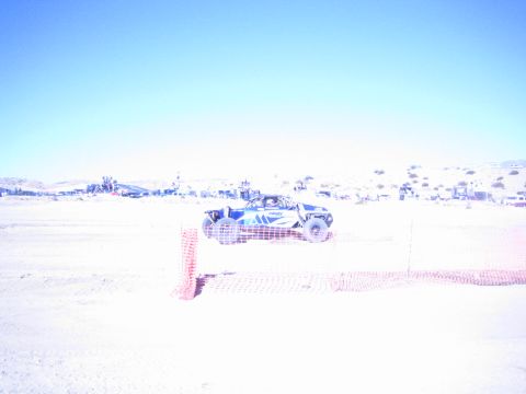 1990 Ford Bronco - blue off-road race car runs a flat desert course behind a low orange fence with hills in the background.