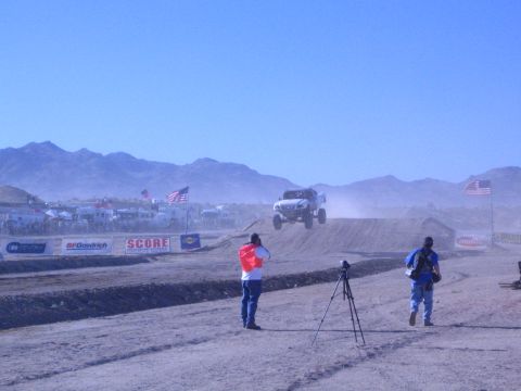 1990 Ford Bronco - photographer and spectator with a tripod watch an off-road truck jump a desert course past banners reading SCORE and BFGoodrich.