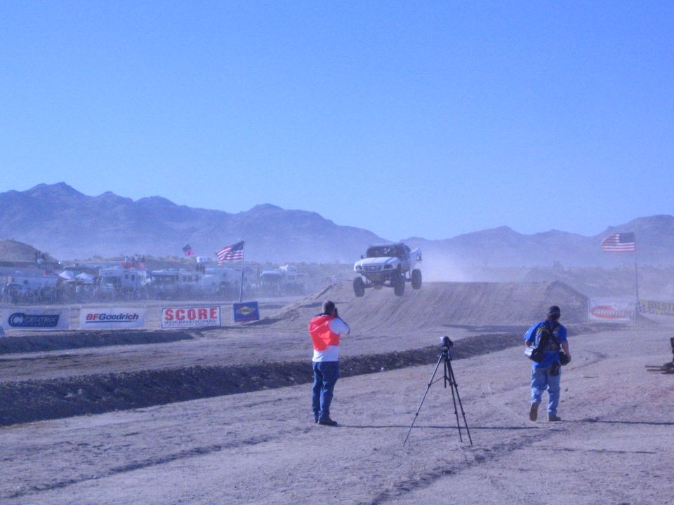 Spectators with camera tripod watch an off-road truck jump on a desert track with US flags and SCORE banners