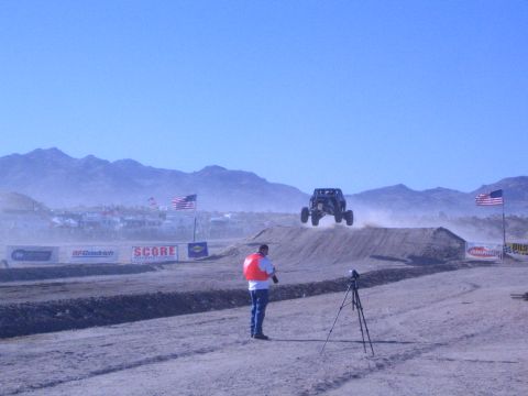1990 Ford Bronco - off-road buggy flies off a dirt jump while a vested man films from a tripod, with US flags and SCORE and BFGoodrich banners lining the cour…