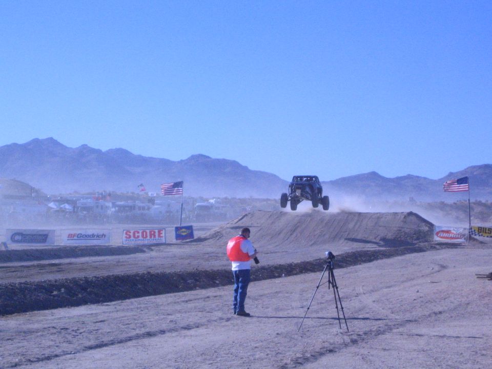 Off-road buggy airborne over a dirt jump as a man films beside US flags and sponsor banners