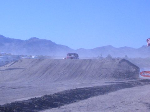 1990 Ford Bronco - dark race vehicle crests a long dirt berm in a dusty desert beside a US flag and a red sponsor banner.