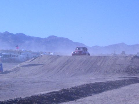 1990 Ford Bronco - dark Baja-style Volkswagen Beetle off-road racer crests a dirt berm in a hazy desert valley with RVs parked behind.