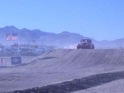 1990 Ford Bronco - dark Baja Bug crosses a dirt berm in a dusty desert beside a US flag and sponsor signage along the course.