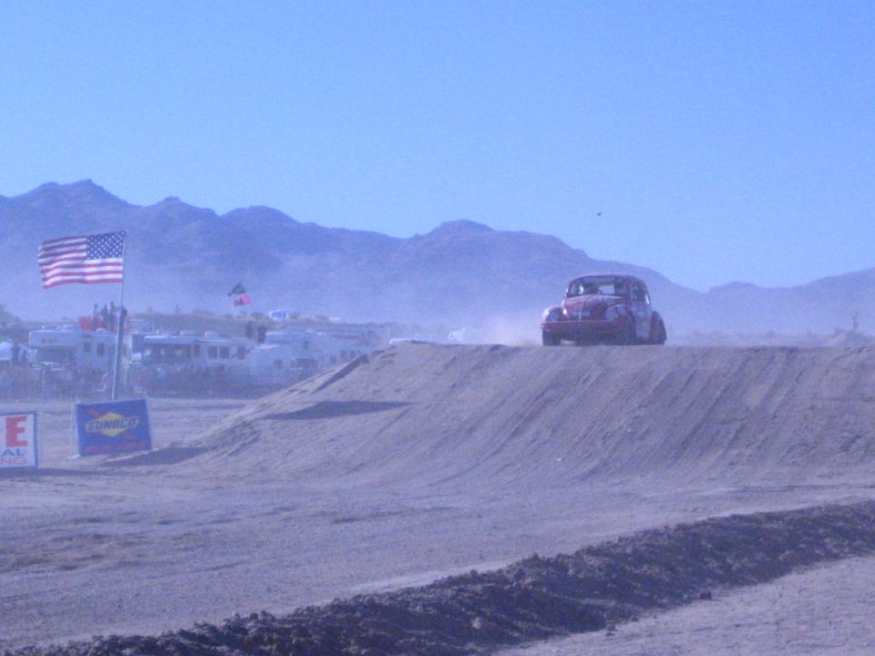 Dark Baja Bug on a dirt berm with a US flag and sponsor signs in a dusty desert