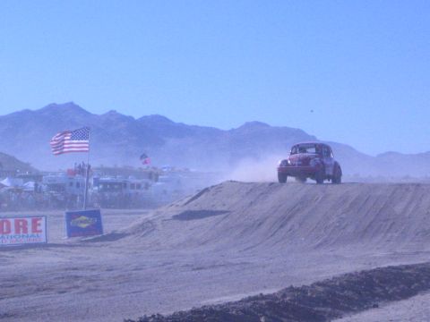 1990 Ford Bronco - dark Baja Bug lifts off a dirt jump in the desert near a US flag and a SCORE International banner.
