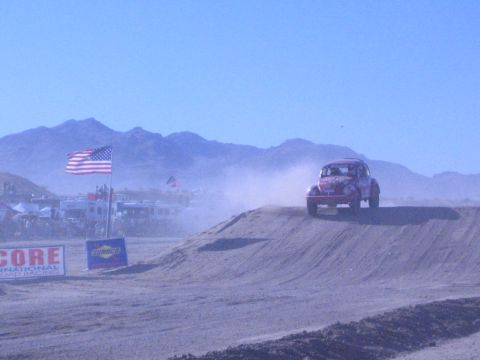 1990 Ford Bronco - red Baja Bug crests a dirt berm trailing dust, with a US flag and SCORE and Sunoco banners along the desert course.