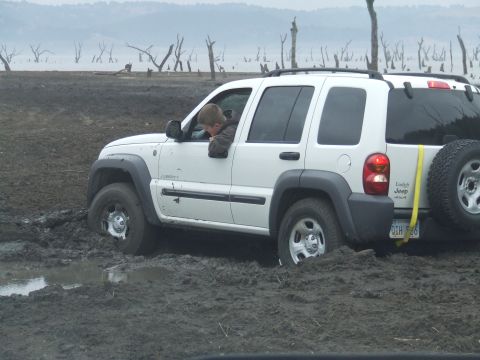 1994 Ford Bronco - A white Jeep Liberty is stuck in thick mud near a body of water with dead trees.