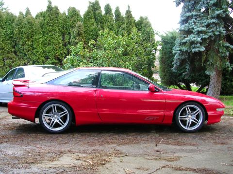 1996 Ford Probe - A red 1996 Ford Probe GT is seen from the side, parked next to another vehicle with trees in the background.