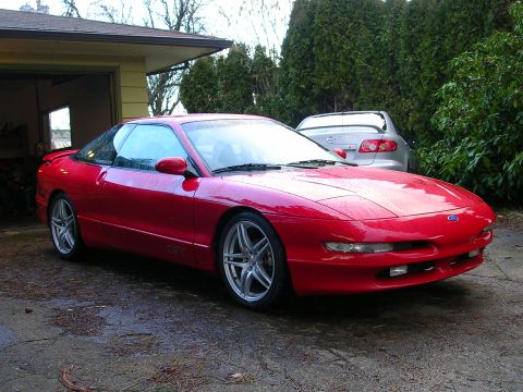 1996 Ford Probe - A red 1996 Ford Probe is parked in a driveway next to a garage, with another vehicle visible behind it.