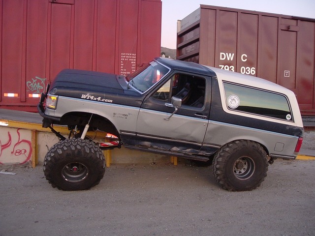 Lifted two-tone Ford Bronco on big mud tires, side view by maroon boxcars