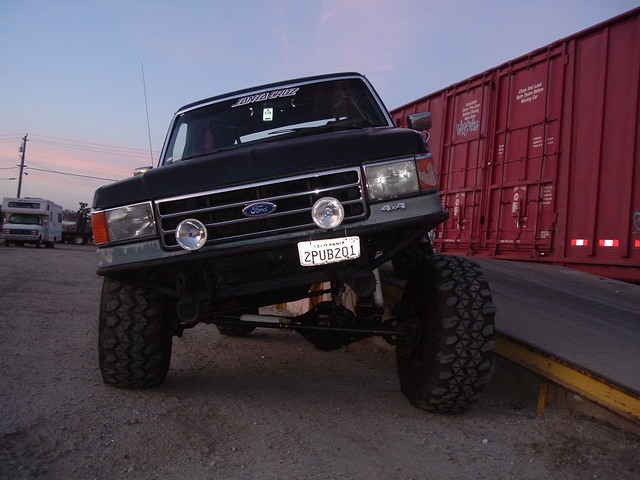 Black lifted Ford Bronco with driving lights and mud tires, front view at dusk beside red railroad boxcars