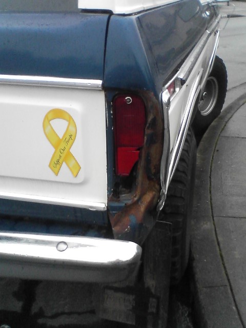 1978 Ford Bronco - Rear view of a blue and white SUV, showing a yellow 'Support Our Troops' ribbon sticker and significant damage to the taillight assembly.