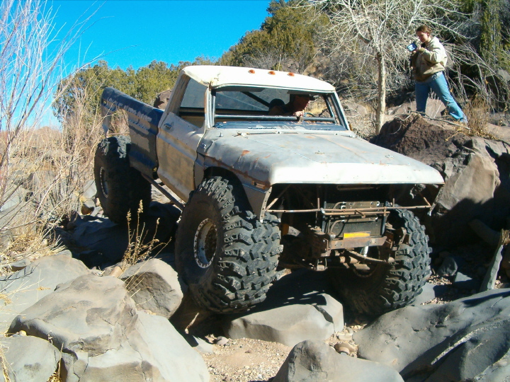 Rusty lifted Ford pickup flexing over dark boulders with spotter standing by