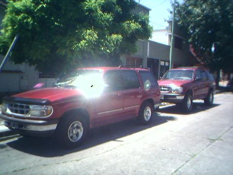 1996 Ford Explorer - Two red 1996 Ford Explorers are parked on a street with trees and buildings in the background.