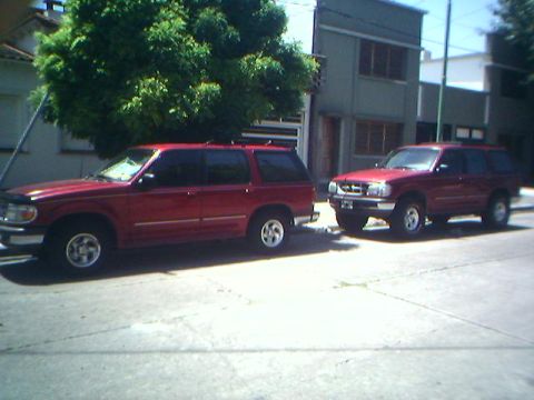 1996 Ford Explorer - Two red Ford Explorer SUVs are parked side-by-side on a street in front of a building.