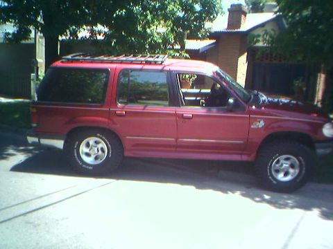 1996 Ford Explorer - A red Ford Explorer SUV is parked on a street, viewed from the side with a roof rack visible.