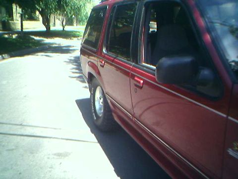 1996 Ford Explorer - The side profile of a red SUV is visible, parked on a sunlit street with trees in the background.