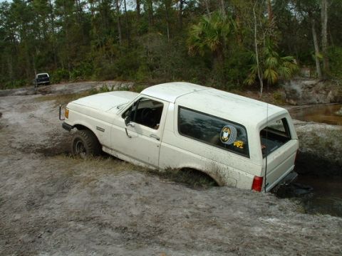 1989 Ford Bronco - A white 1989 Ford Bronco navigates a muddy trail, with another vehicle visible in the background.