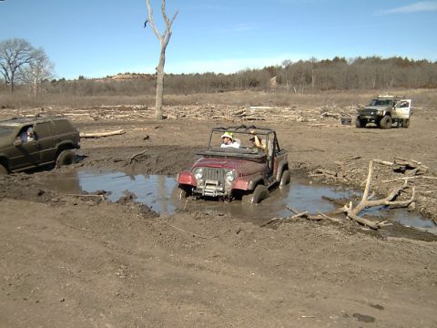 1998 Jeep TJ - A red 1998 Jeep TJ is stuck in a muddy puddle while two other off-road vehicles are visible in the background.