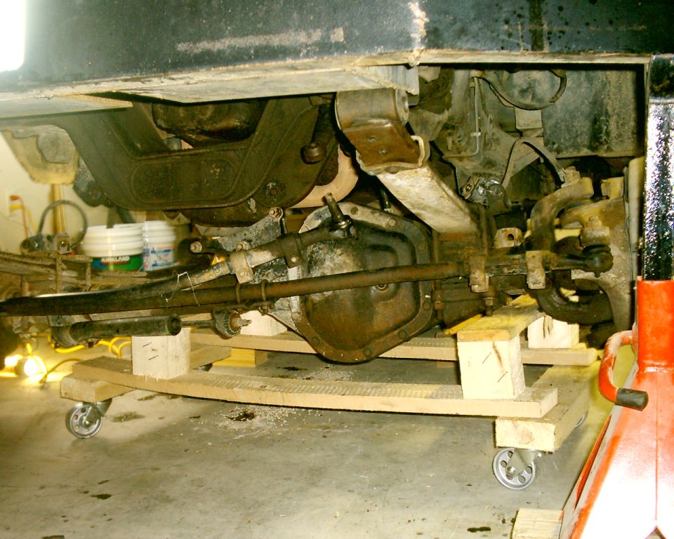 Under a Bronco: front solid axle, differential and steering linkage supported on wood blocks and a rolling dolly in a garage.