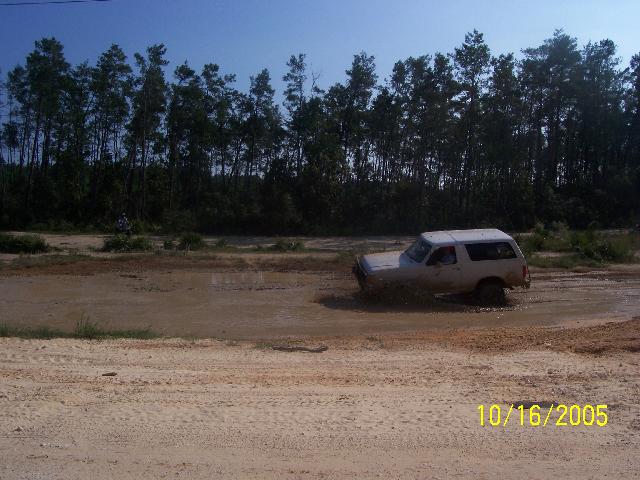 1989 Ford Bronco driving through a muddy puddle with trees in the background