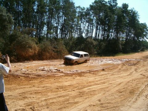 1989 Ford Bronco - A white SUV is stuck in a deep mud puddle on a dirt road, with trees lining the path.