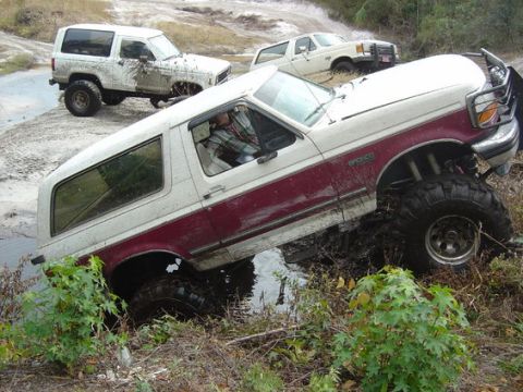 1989 Ford Bronco - Several SUVs navigate a muddy off-road course, with one Bronco deeply mired in the water.