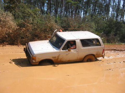 1989 Ford Bronco - This 1989 Ford Bronco is deeply stuck in a muddy off-road trail, with its driver visible behind the wheel.