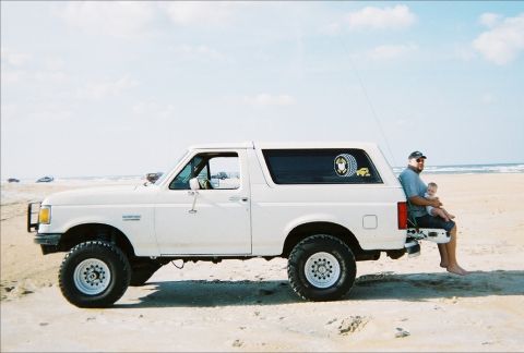 1989 Ford Bronco - A man and baby relax on the tailgate of a white Ford Bronco on a sunny beach day.