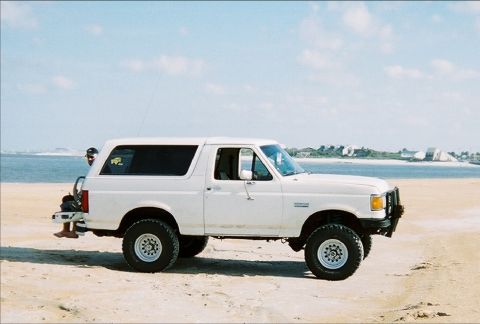 1989 Ford Bronco - A white 1989 Ford Bronco is parked on a sandy beach with the ocean in the background.