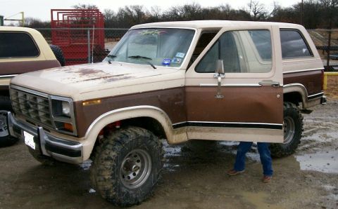 1993 Ford Bronco - A two-tone tan and brown early-1980s Ford Bronco full-size SUV shown in 3/4 front view, lifted on large mud-terrain tires with rust on the…