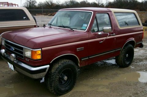 1993 Ford Bronco - Three-quarter front view of a maroon Ford Bronco with a tan removable hardtop, parked in a muddy gravel lot beside a chain-link fence. It w…