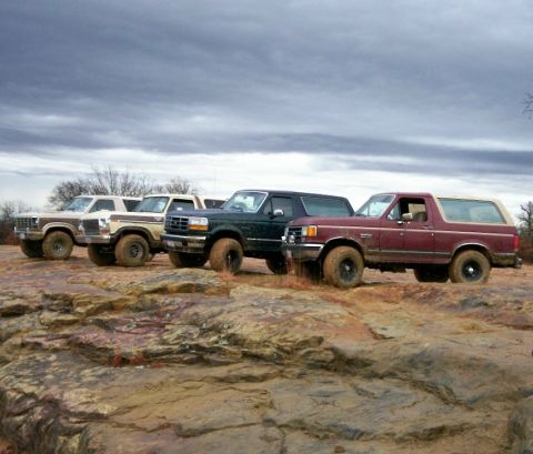 1993 Ford Bronco - Four full-size Ford Bronco SUVs lined up nose-to-tail on a slab of red sandstone ledge under heavy gray storm clouds, all riding on mud-cak…
