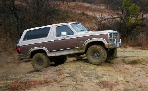 1993 Ford Bronco - Side view of a two-tone brown and cream 1980s Ford Bronco crawling up a wet slickrock ledge on a winter trail. It runs mud-terrain tires on…