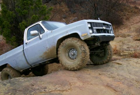 1993 Ford Bronco - A white square-body Chevrolet regular-cab 4x4 pickup crawls up a steep rock ledge, front end lifted and mud smeared down the doors and bed.…