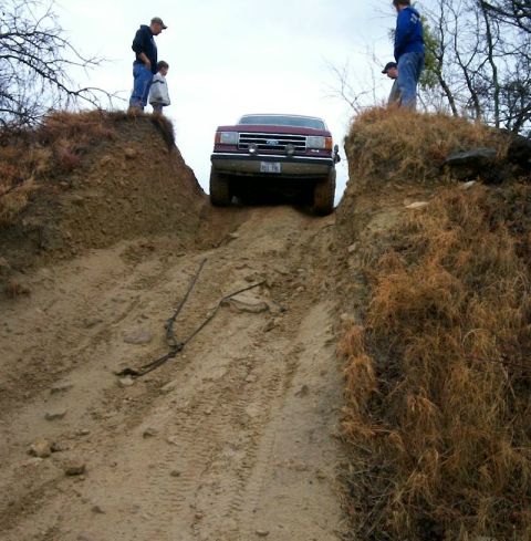 1993 Ford Bronco - Shot from the bottom of a steep rutted dirt hill, a dark red Ford truck with a chrome grille, blue Ford oval and grille-mounted driving lig…