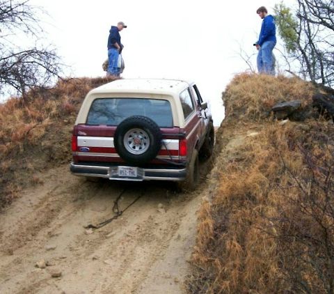 1993 Ford Bronco - Rear view of a two-tone dark red and cream full-size Ford Bronco climbing a steep rutted dirt chute, with a swing-away tire carrier holding…
