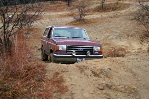 1993 Ford Bronco - A maroon Ford Bronco with a tan top climbs out of a dirt washout on a bare, eroded off-road trail, its front wheels dug into the rut. Round…