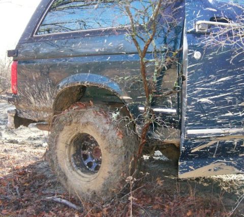 1993 Ford Bronco - Close rear-quarter view of a dark blue full-size Ford SUV bogged in mud, its open driver's door and body sides flecked with slung mud spray…