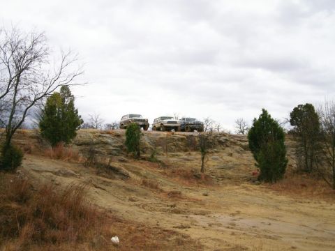 1993 Ford Bronco - Three lifted 4x4 trucks, two light-colored SUVs and a dark blue one, parked side by side on a rock ledge at the top of a sandy off-road hil…