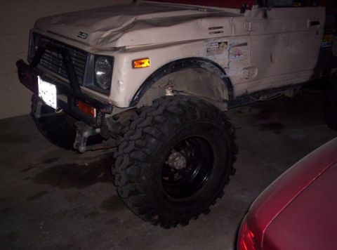 1987 Suzuki Samurai - A beige 1987 Suzuki Samurai with significant body damage and oversized tires is parked indoors.